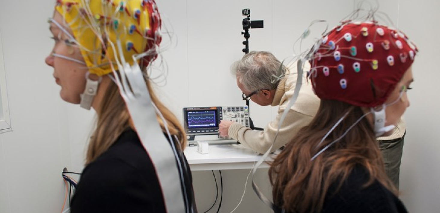 Två studenter med EEG-elektrodmössor på huvudet. I bakgrunden en forskare. Foto: Jens Olof Lasthein